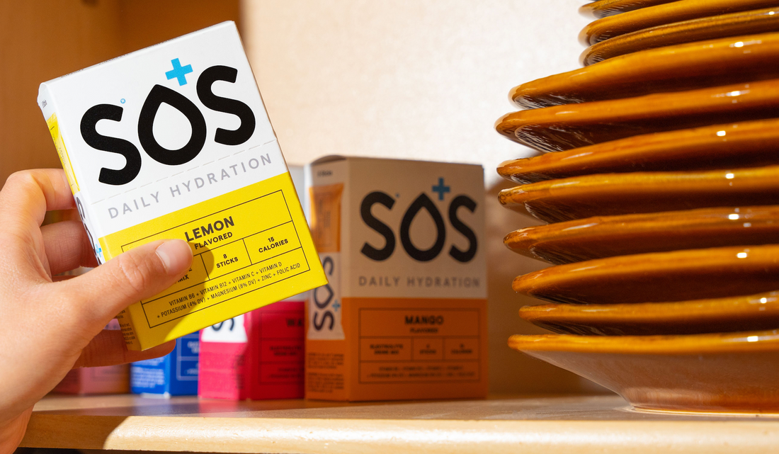 Hand reaching for a box of SOS Daily Hydration Lemon electrolyte drink mix on a kitchen shelf next to stacked plates and other SOS flavors.